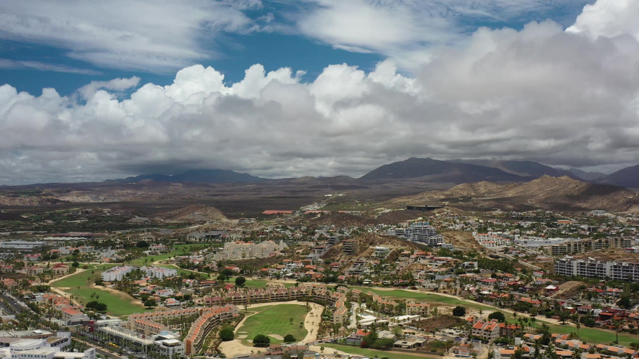 Los Cabos aerial frame with clustered buildings, curved roads, bright green open areas, layered hills, distant mountains, and no visible people.