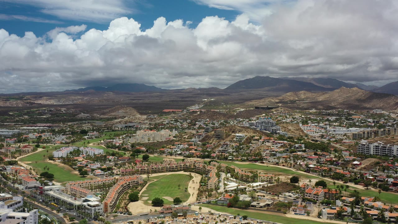 Wide aerial view of homes, roads, green fairways, dry hills, mountains, and clouds.