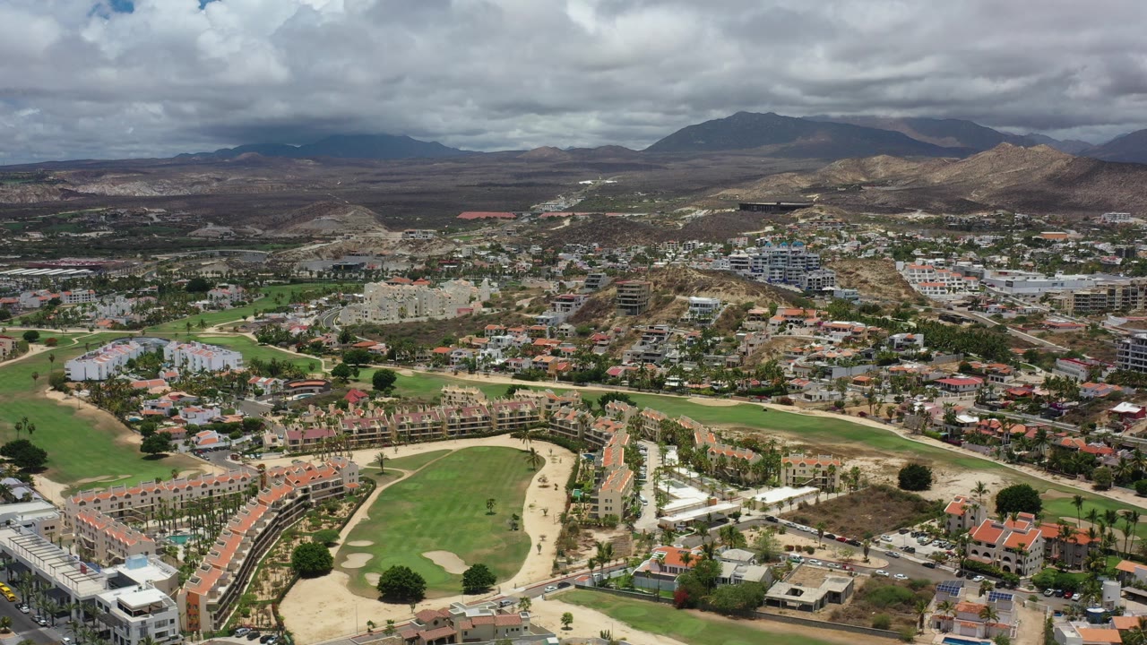 Aerial view of suburban homes, golf greens, roads, hills, and cloudy mountains.