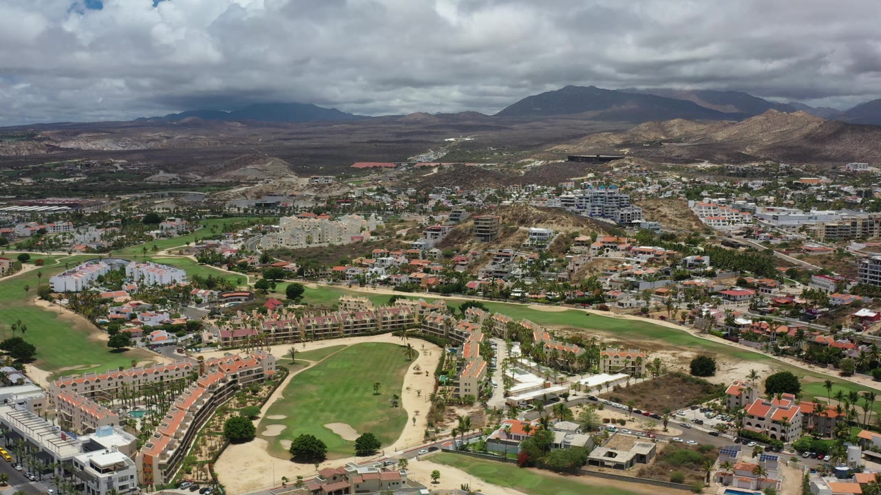 Los Cabos aerial view one second into the clip, with residential streets and fairways filling the foreground and a darker cloud band over the mountain ridge, no visible people.