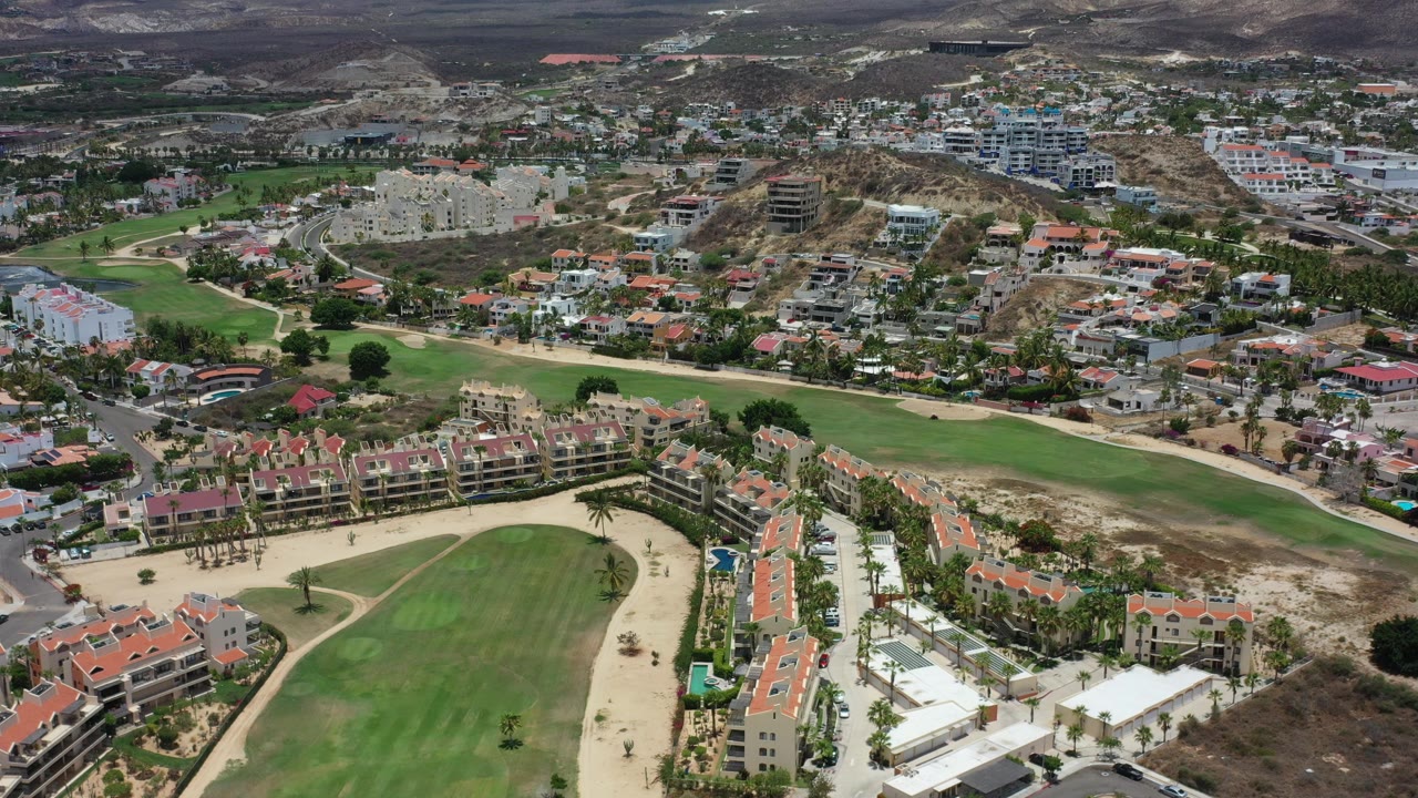 Wide aerial view over Los Cabos, Mexico, showing a green golf fairway, red-roofed residential buildings, roads, dry hills, and no visible people at the start of the clip.