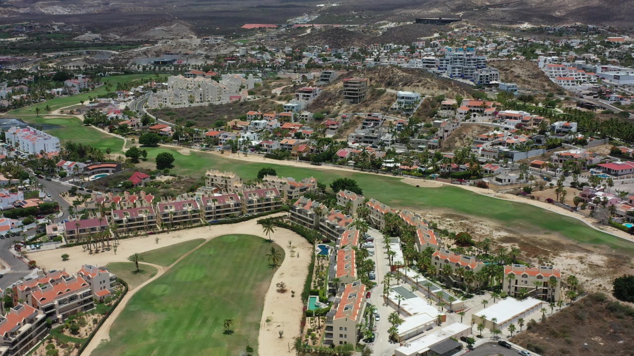 Aerial frame over Los Cabos, Mexico, with the fairway sweeping across the lower frame, clustered homes and apartment blocks beyond, and no visible people.