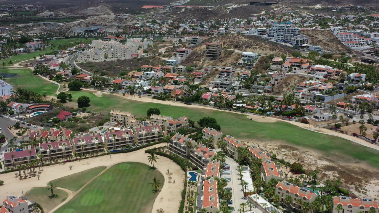 Aerial Los Cabos frame showing a wide green fairway cutting through residential buildings, with hillside construction and dry terrain behind, and no visible people.