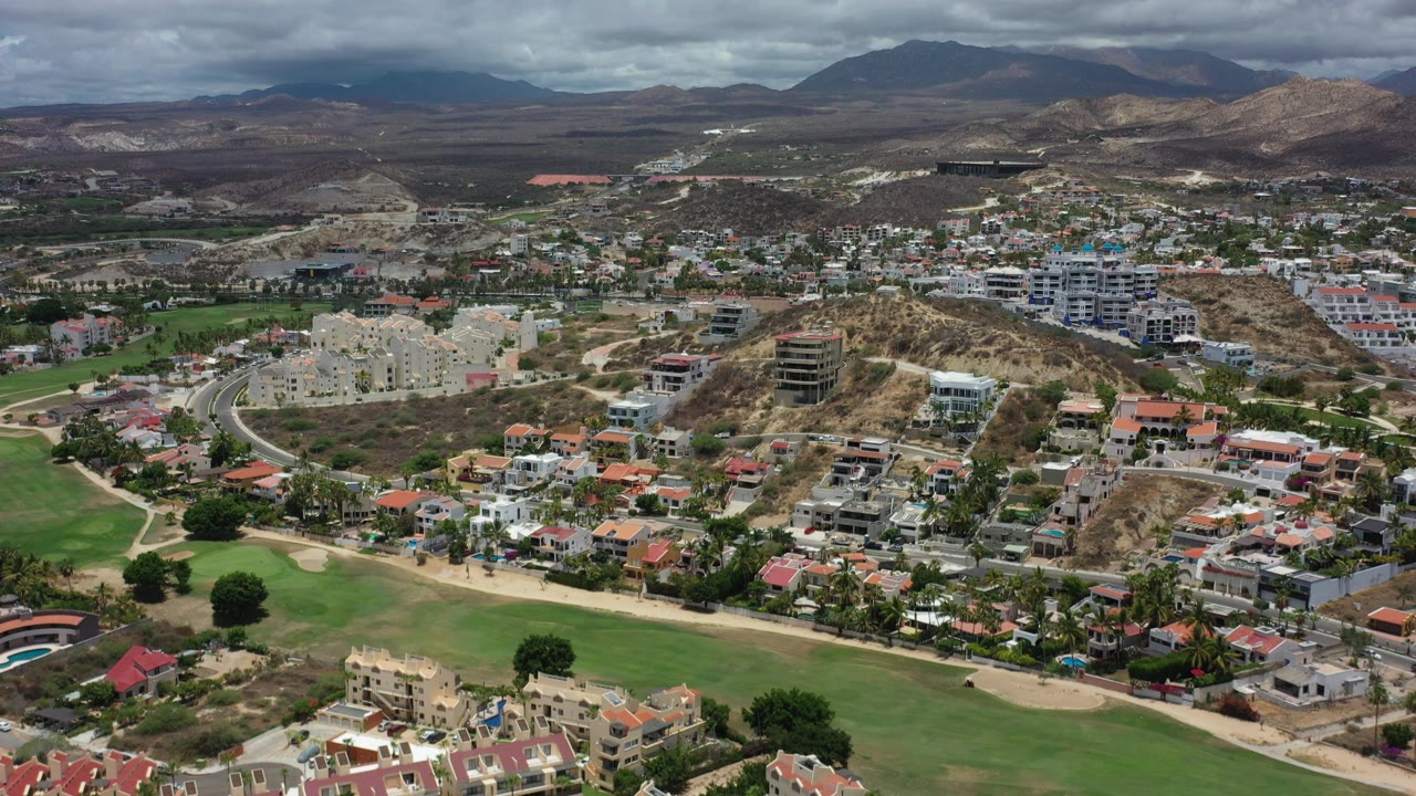 Opening aerial frame over Los Cabos, Mexico, with green fairway areas in the foreground, hillside residential buildings in the middle distance, dark mountains on the horizon, and no visible people.