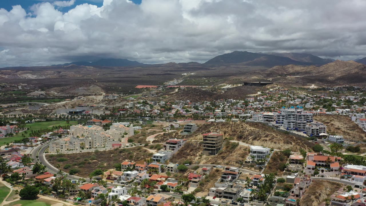 Los Cabos aerial view one second in, showing a curved road at lower left, clusters of homes, tan hills, mountains on the horizon, cloud shadows, and no visible people.