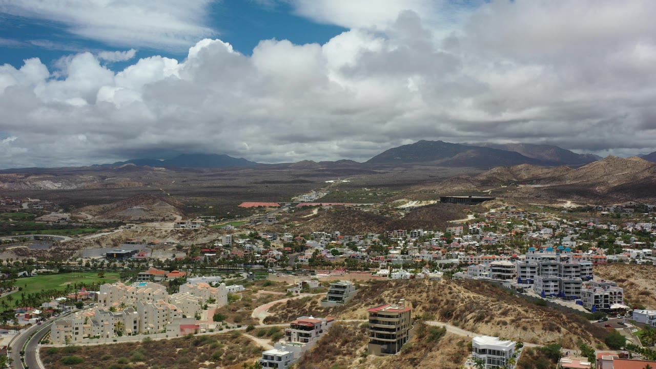 Aerial view over Los Cabos, Mexico, with apartment blocks, roads, dry hills, distant mountains, heavy clouds, and no visible people at the start of the clip.
