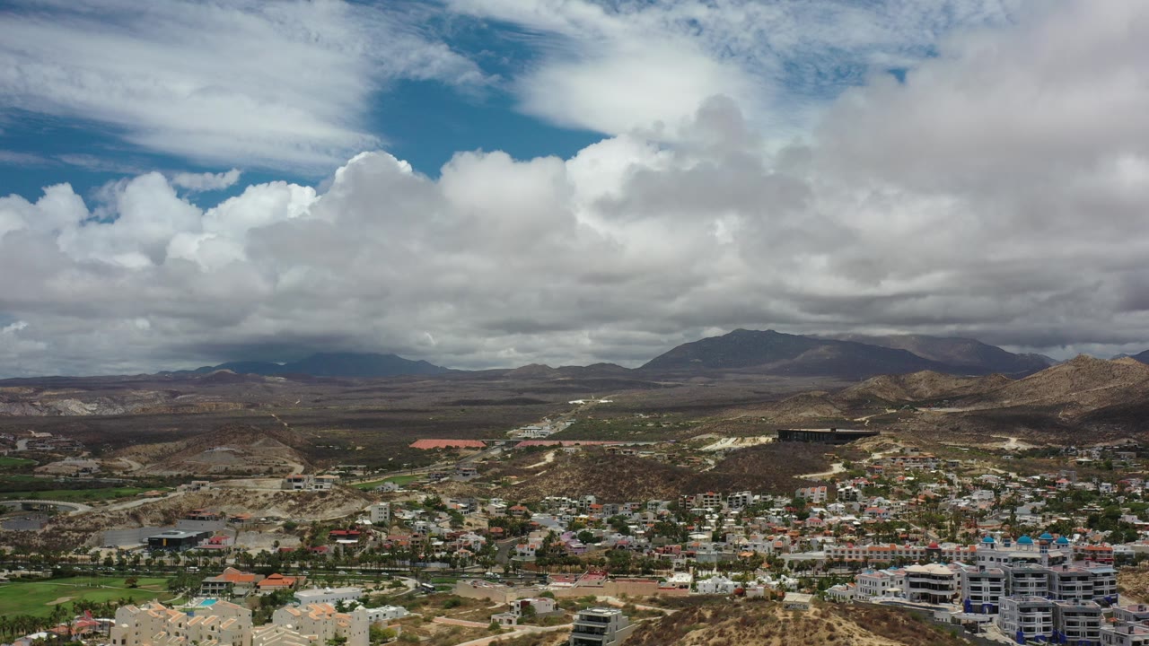 Opening view of Los Cabos, Mexico, with low-rise buildings below dry hills and distant mountains under large layered clouds, with no visible people.