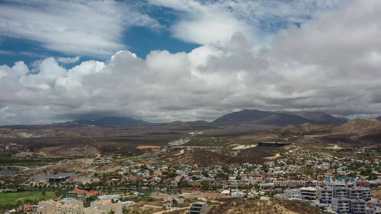 Los Cabos appears from above with clustered town buildings in the lower frame, brown hills beyond, and thick white-gray clouds crossing the mountain horizon, with no visible people.
