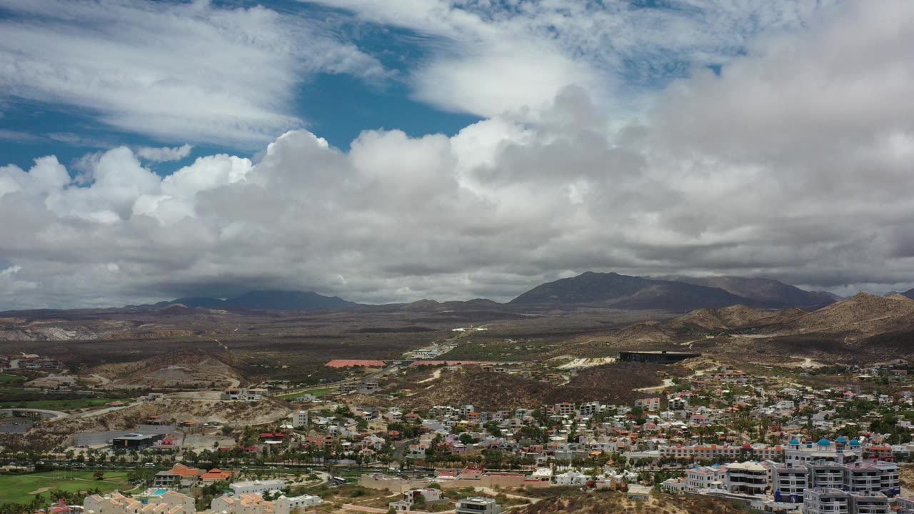 Wide aerial composition of clouds, mountains, hills, and urban rooftops.