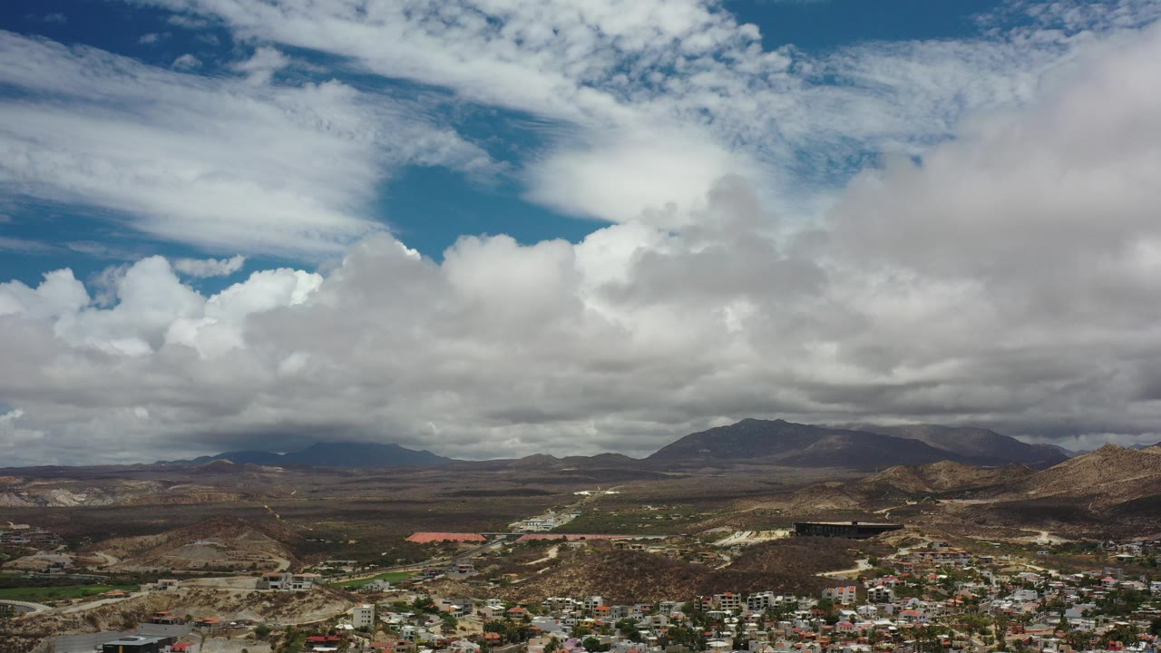 Opening frame of Los Cabos, Mexico, showing low buildings at the bottom, dry hills, distant mountains, and layered white clouds with no visible people.