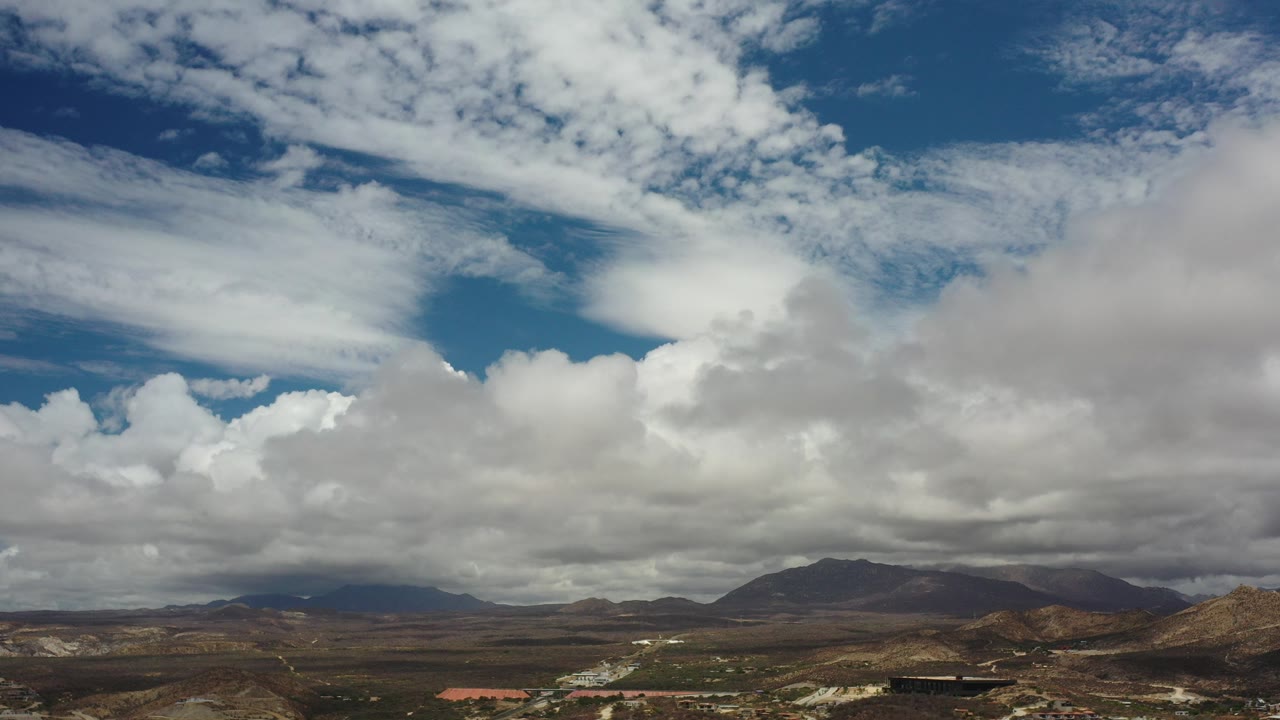 Opening frame of Los Cabos, Mexico with a broad blue sky, thick white and gray clouds, a low desert horizon, distant mountains, and no visible people.