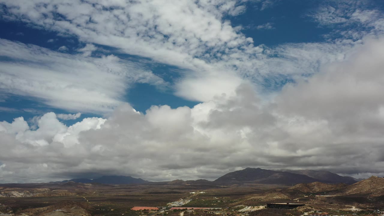 Los Cabos sky view with a bright blue opening between layered clouds, dark hills along the horizon, sparse development below, and no visible people.