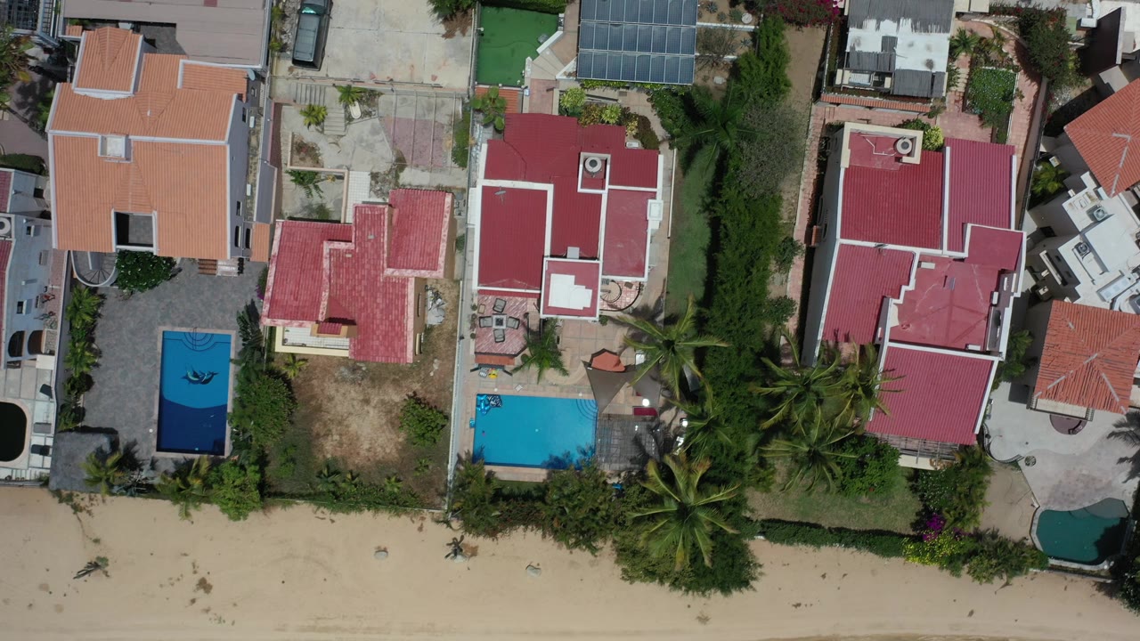 Top-down view in Los Cabos, Mexico showing red and orange roofed homes, two blue swimming pools, palms, driveways, and a sandy beach edge with no visible people.