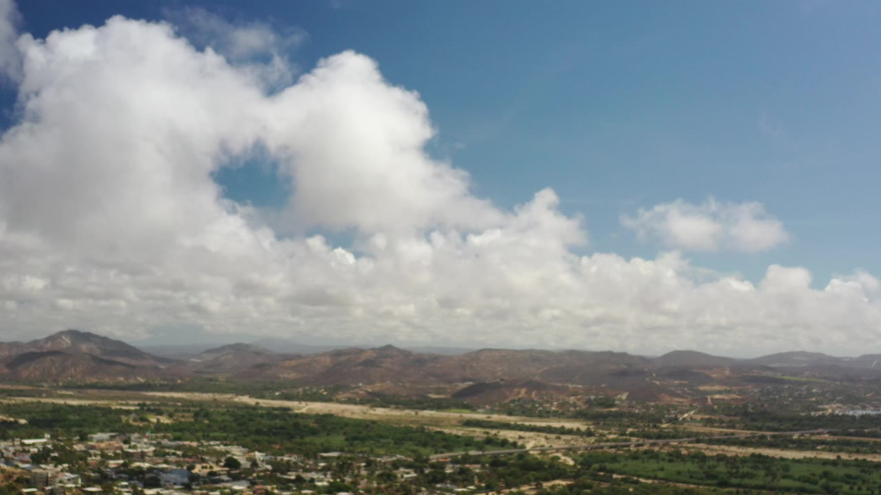 Aerial view of city edge, green land, mountains, clouds, and blue sky.