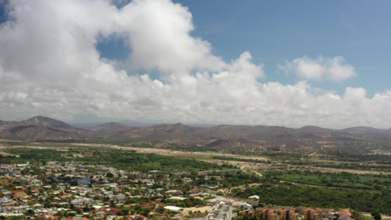 Wide aerial view of a neighborhood below brown mountains and large clouds.