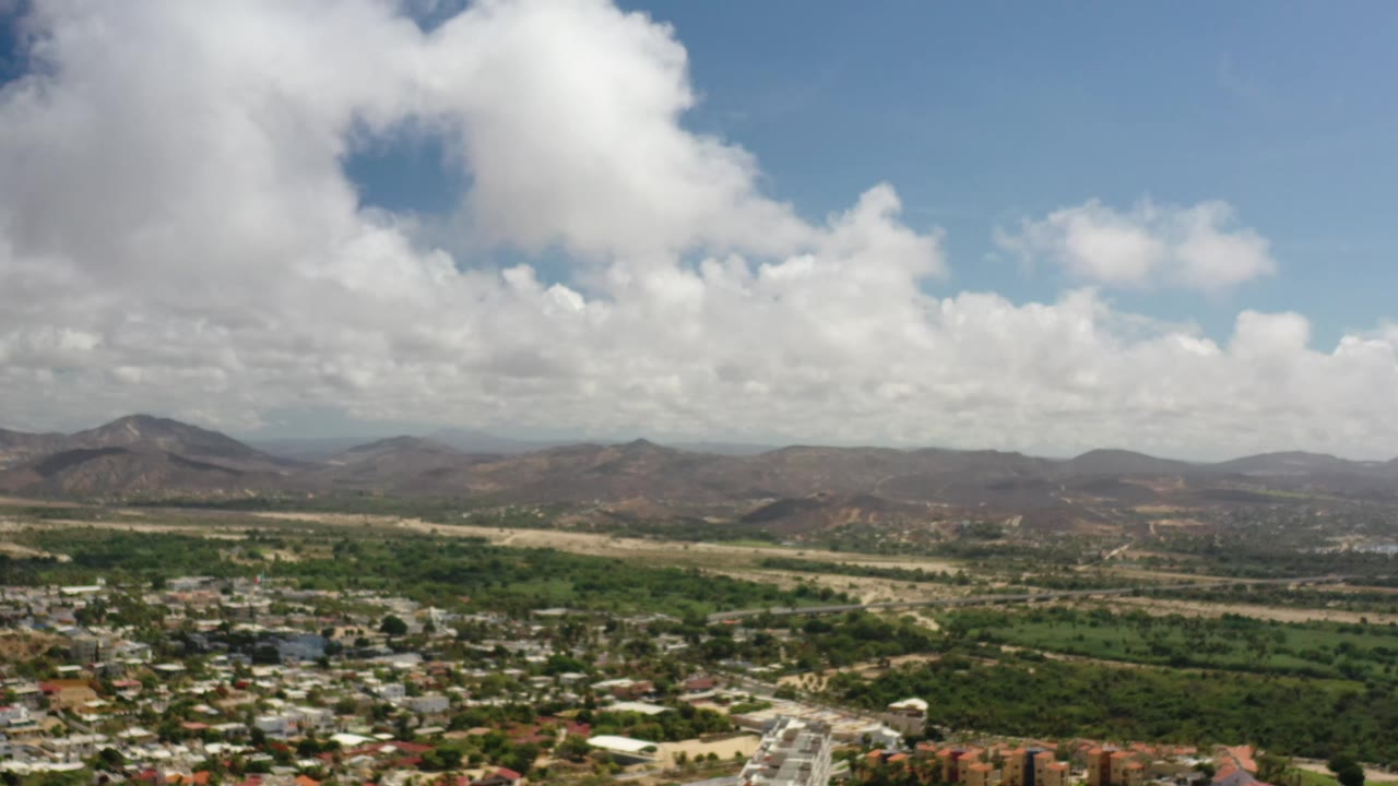 Early Los Cabos frame with clustered rooftops low in the image, a green belt through the midground, dry terrain beyond it, and mountain ridges under bright clouds with no visible people.