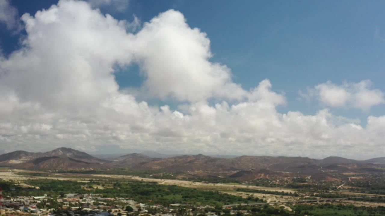 Opening frame of Los Cabos, Mexico with large white clouds over brown hills, green valley patches, and no visible people.