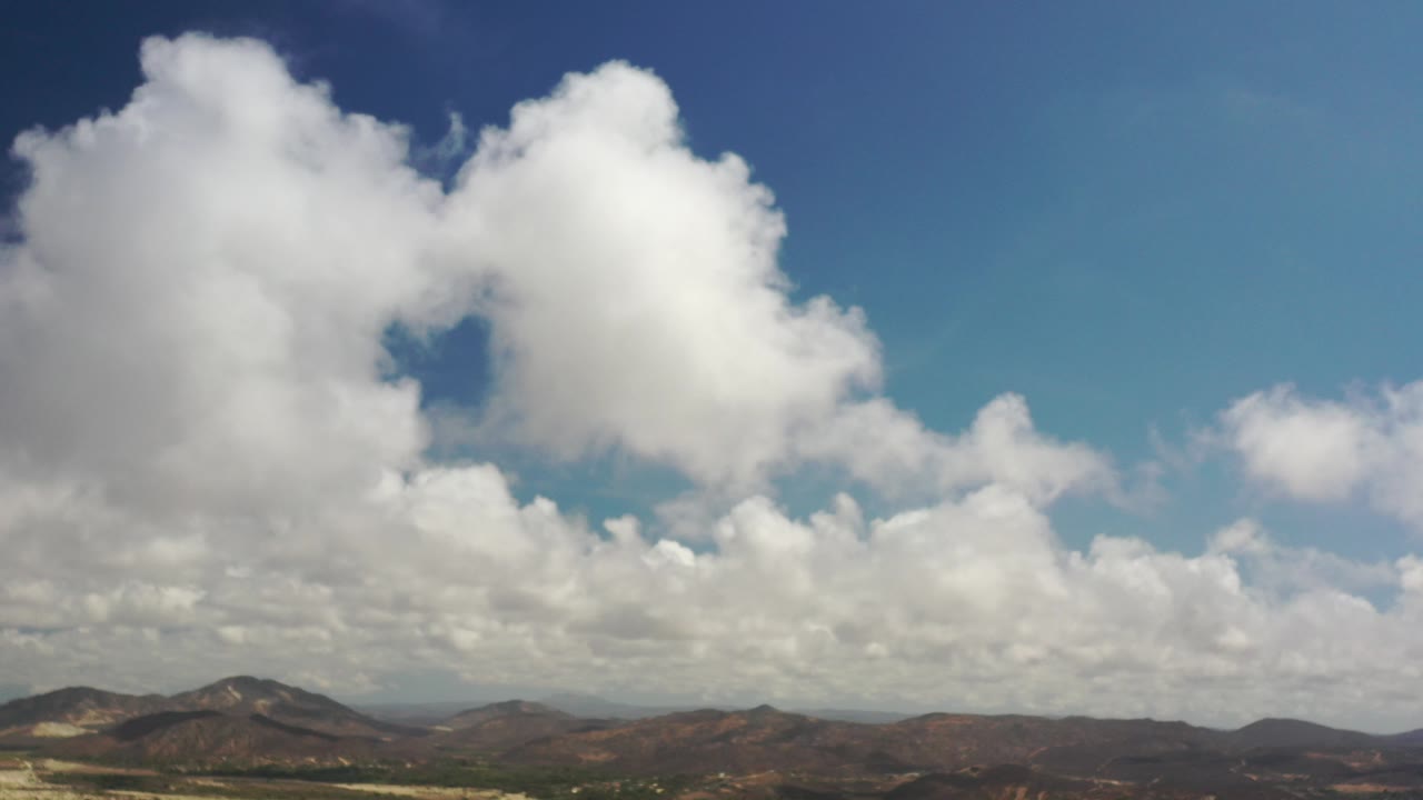 Wide Los Cabos aerial view with large white clouds filling the blue sky above distant brown hills and no visible people.