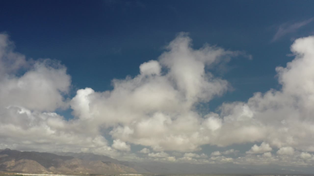 Opening frame of Los Cabos, Mexico, showing a wide blue sky with bulky white clouds over a low mountain horizon and no visible people.