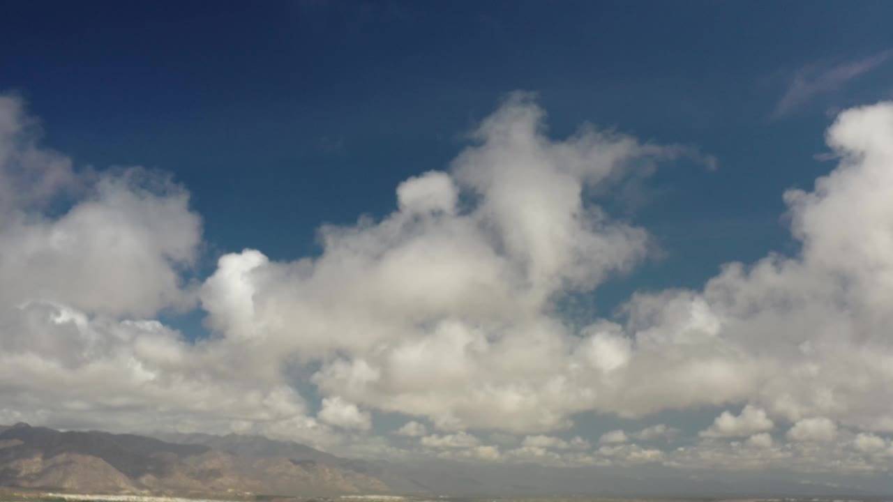 A broad Los Cabos sky view shows clustered white clouds above hazy mountains, with a narrow band of city detail near the bottom and no visible people.
