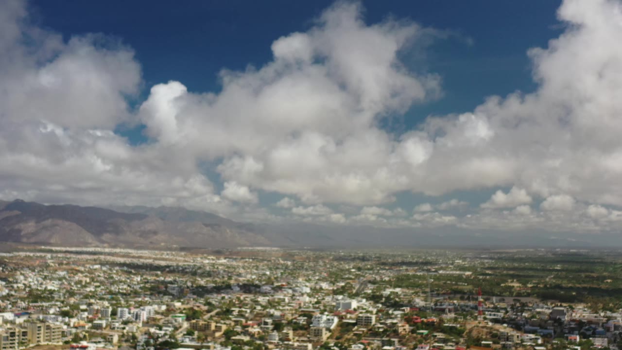 Early frame of Los Cabos, Mexico, showing urban blocks across the lower frame, a hazy mountain ridge on the horizon, large white clouds, and no visible people.