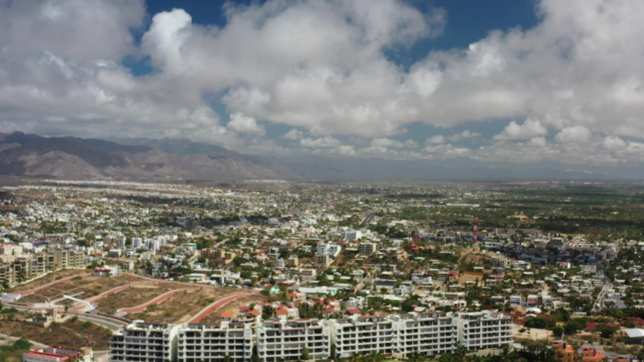 Opening aerial frame over Los Cabos, Mexico, showing apartment blocks, city streets, distant brown mountains, and large white clouds, with no visible people.