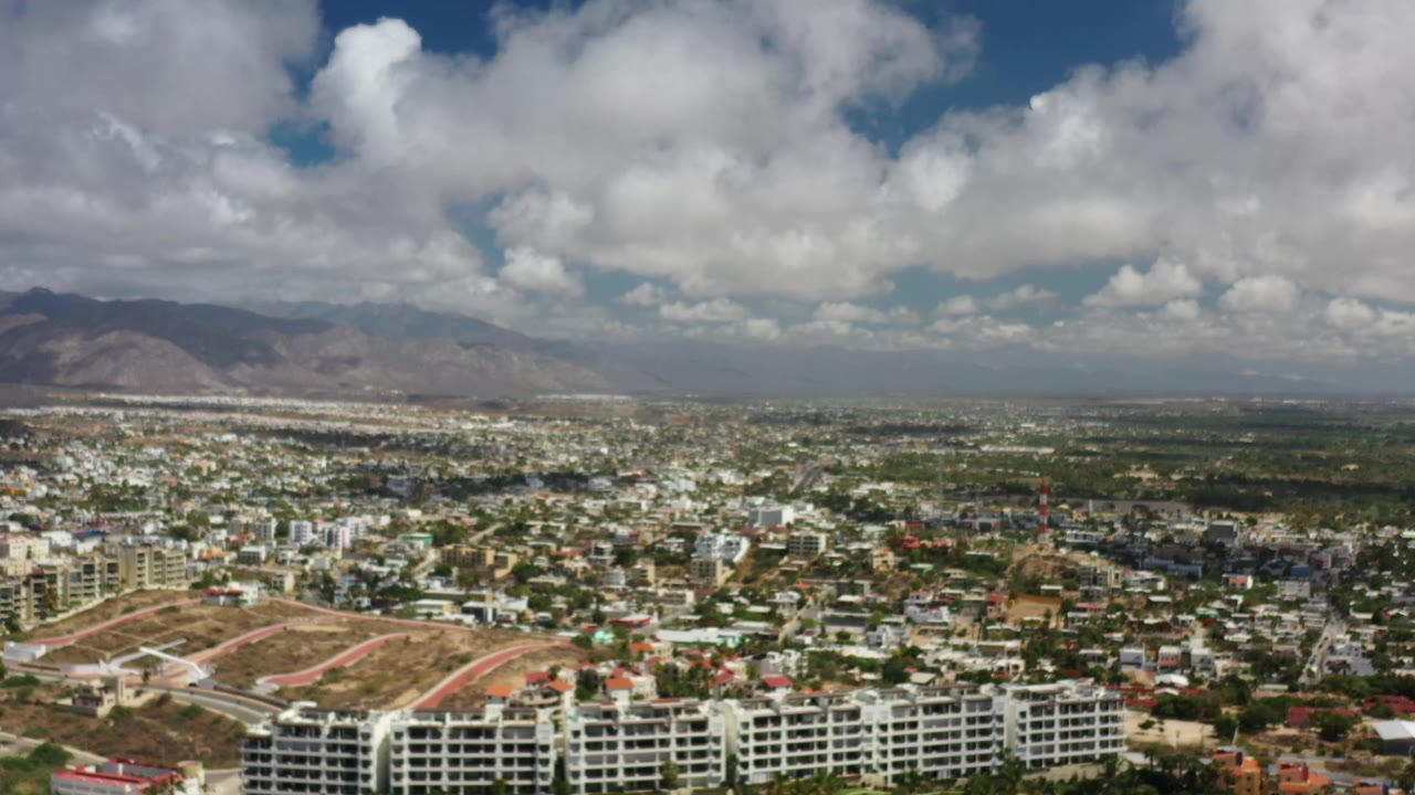 Second aerial frame over Los Cabos with modern buildings in the lower frame, a broad urban spread in the midground, mountains on the horizon, and no visible people.