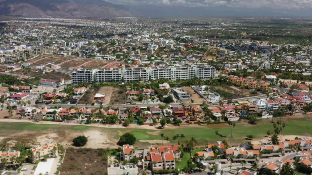 Aerial view across Los Cabos with clustered homes in the foreground, green fairway-like ground, modern white buildings, and hazy hills beyond, with no visible people.