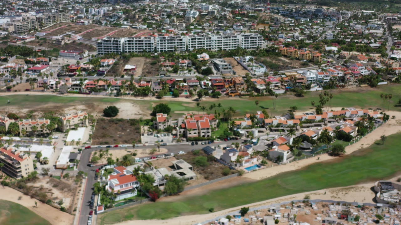 Aerial view over Los Cabos showing red-roofed homes, a green fairway, pale dirt roads, and dense city blocks in the distance, with no visible people.