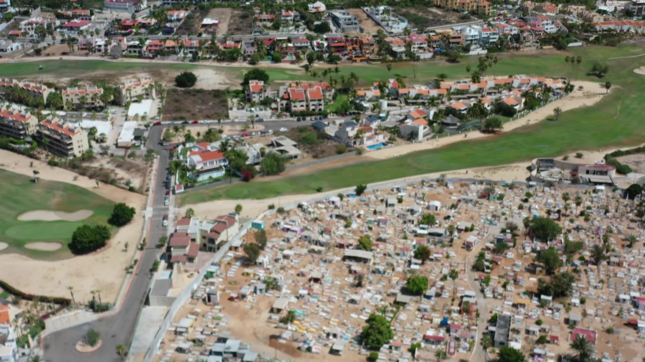 Aerial frame over Los Cabos with the cemetery centered lower in view, a pale road cutting upward, green fairways and bunkers to the left, and red-roof homes in the background.