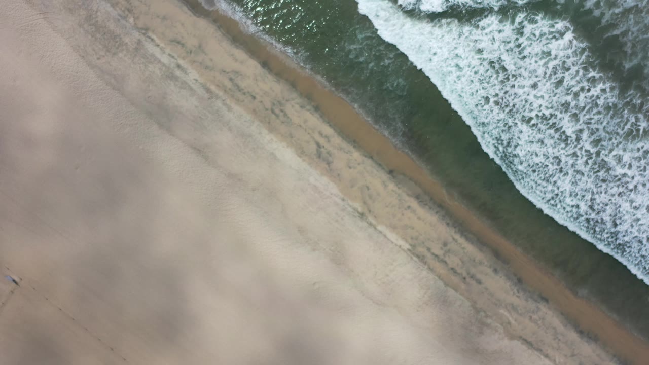Aerial frame over Los Cabos sand where a narrow wet strip separates pale beach from foamy water near the top edge, with no visible people.