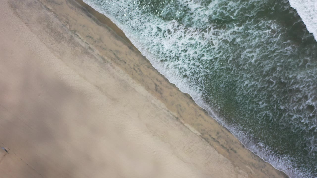 Overhead view in Los Cabos, Mexico, with white surf entering from the upper right and tan beach sand filling the left side, no visible people.