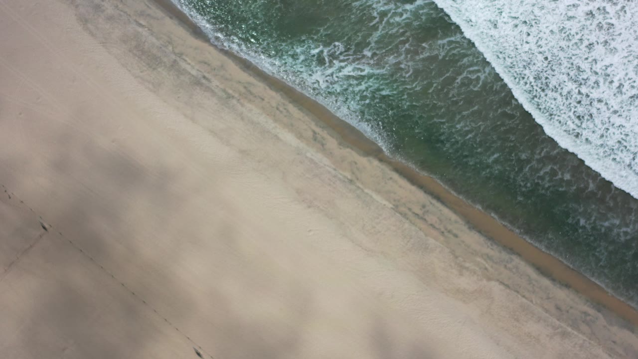Overhead view of a Los Cabos beach where green water and white foam meet tan sand, with soft shadow patches on the dry beach and no visible people.