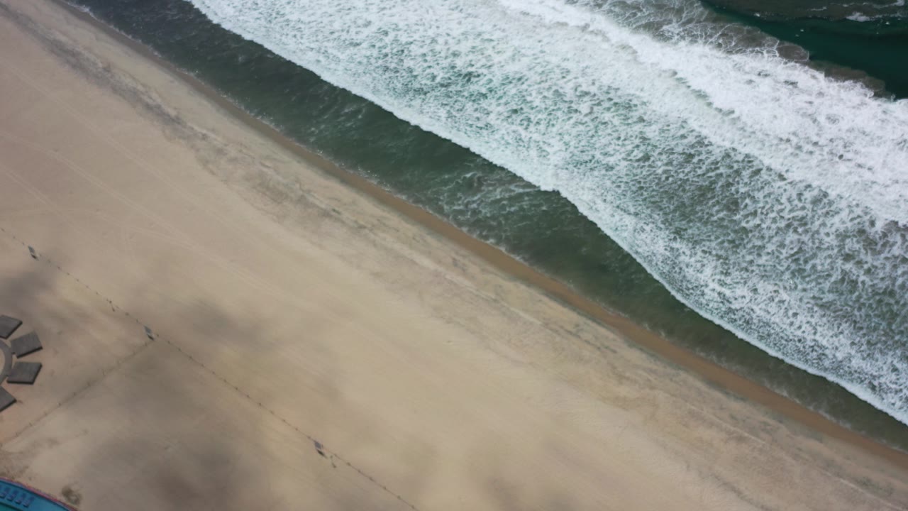 Overhead view in Los Cabos, Mexico, with white surf entering from the upper right across tan sand and no visible people.