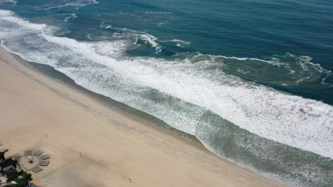 Aerial view over Los Cabos, Mexico, with white surf rolling diagonally onto tan sand and dark blue water beyond, no visible people.