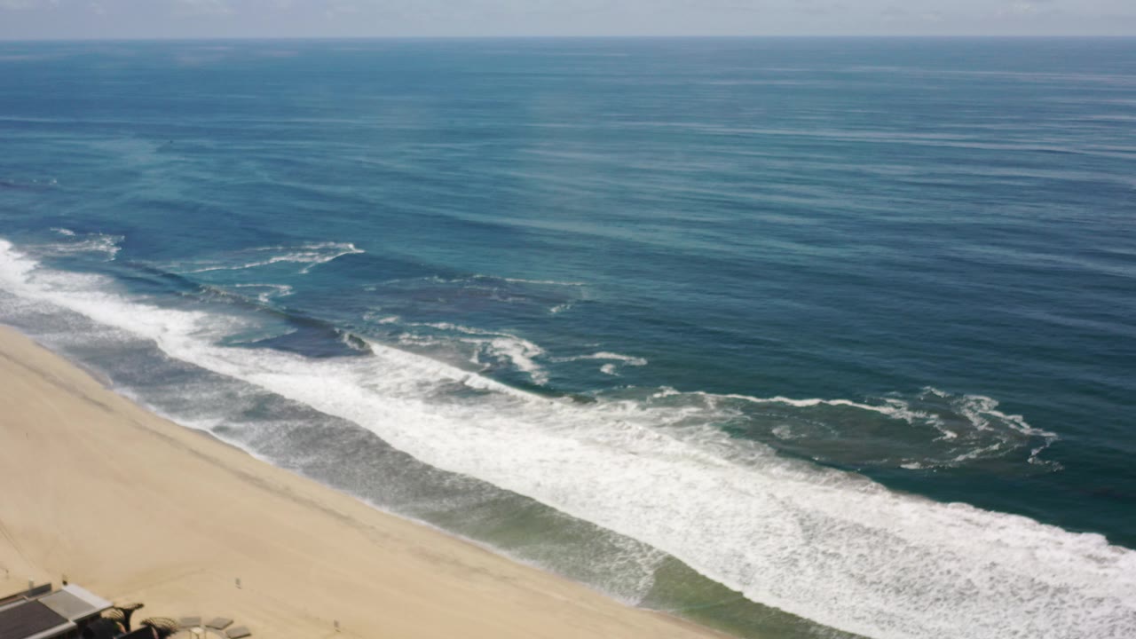 Opening aerial frame over Los Cabos, Mexico, with pale sand at lower left, white surf crossing the shoreline, blue ocean beyond, and no visible people.
