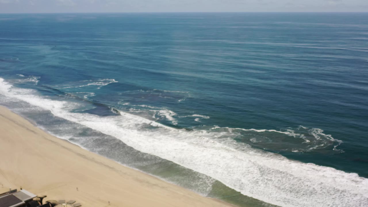 Aerial shoreline view in Los Cabos, Mexico, with a wide white wave band near the beach, darker reef-like patches under the water, and no visible people.