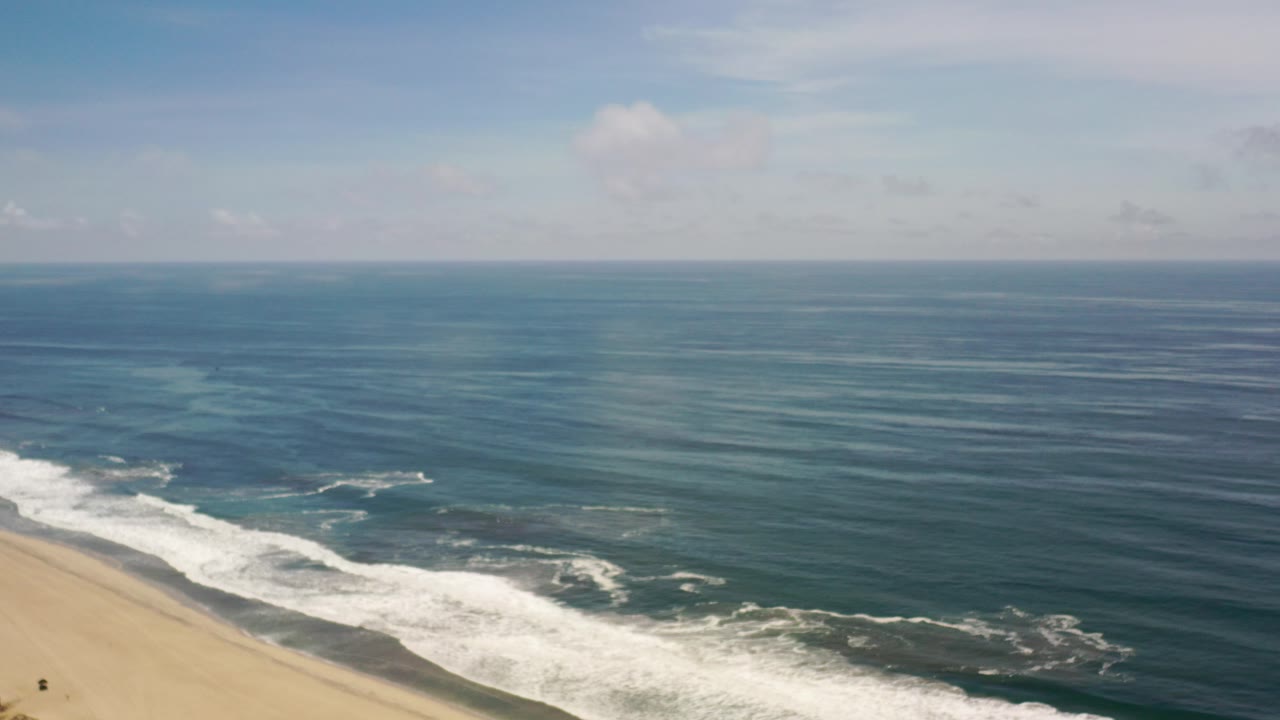 Wide aerial view in Los Cabos, Mexico with tan beach at lower left, white surf rolling along the shore, blue-green ocean bands, pale sky, and no visible people.