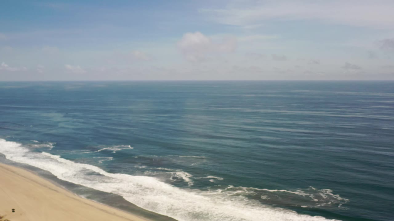 Los Cabos coastline seen from above with foamy waves crossing the lower left, darker offshore water stretching to the horizon, soft cloud cover, and no visible people.