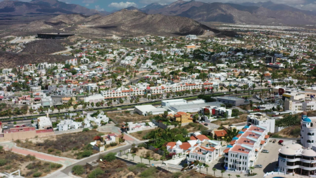 Aerial view of houses, a hotel-style building, pool, desert lots, and mountains.