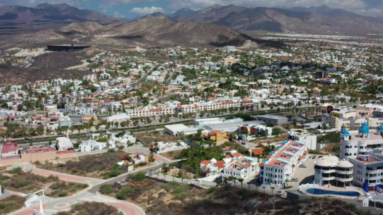 A wide Los Cabos aerial holds on white buildings, scattered greenery, dry open land, distant mountains, and no visible people.