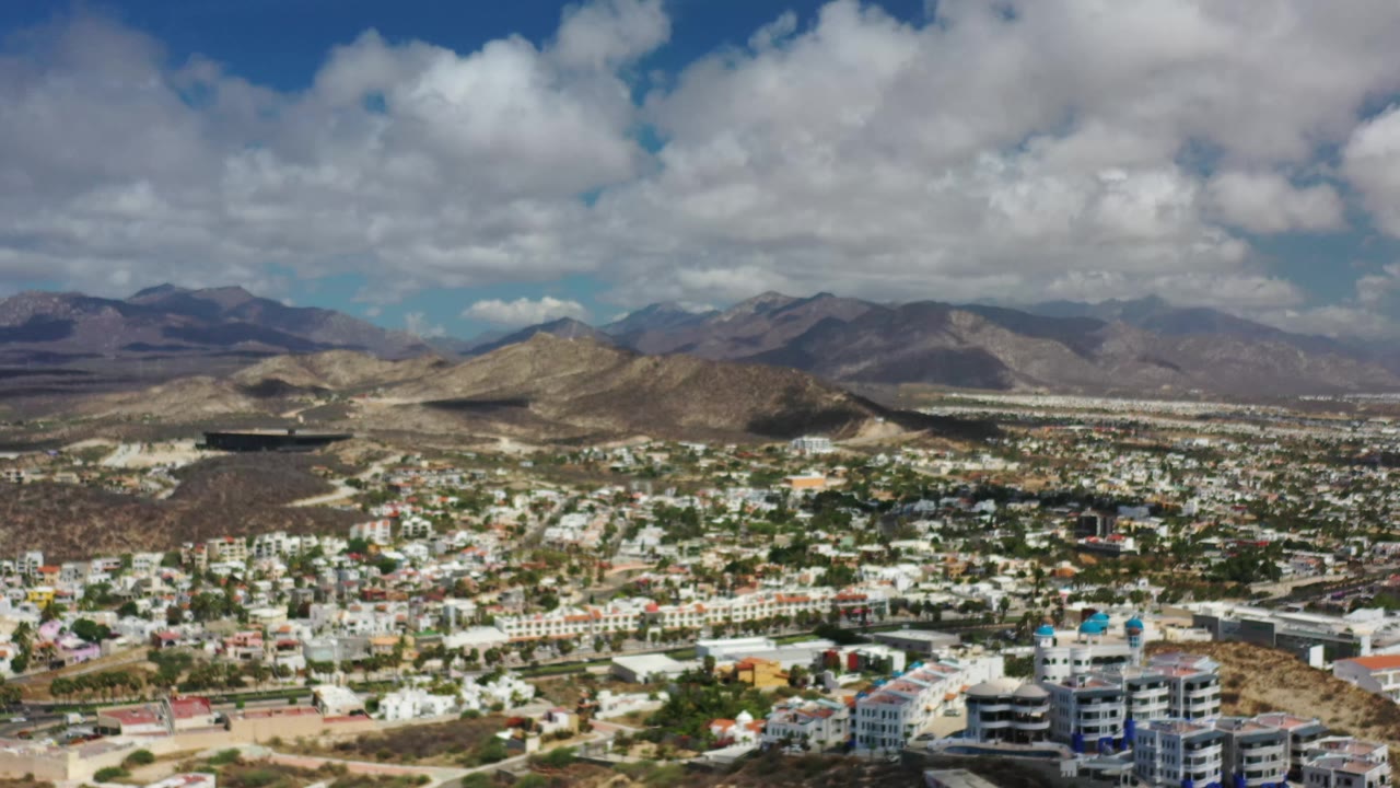 Los Cabos aerial view one second in, showing dense low buildings below arid hills, distant mountain ridges, a blue sky, and no visible people.