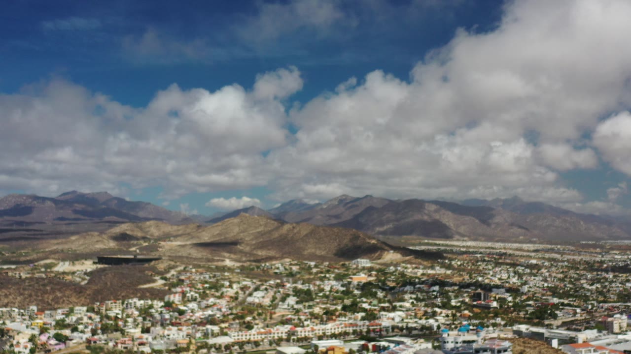 Wide aerial view over Los Cabos, Mexico, with low city buildings at the bottom, tan desert hills, distant mountains, large white clouds, and no visible people.