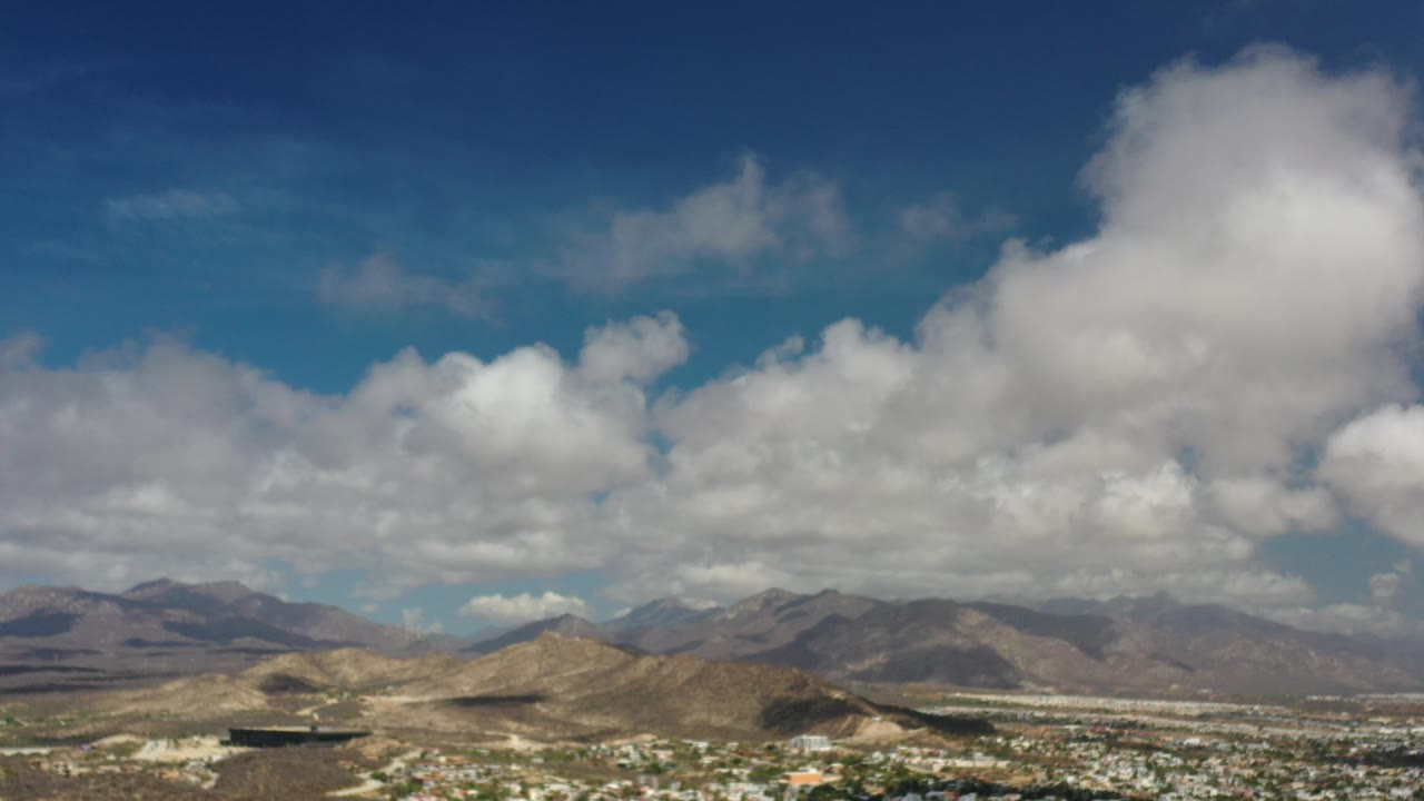 Opening frame of Los Cabos, Mexico with a deep blue sky, broad white clouds, dry mountains on the horizon, distant town detail, and no visible people.