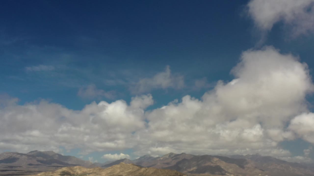 Early frame showing a large pale cloud near the upper center over Los Cabos mountains, with blue sky filling most of the image and no visible people.