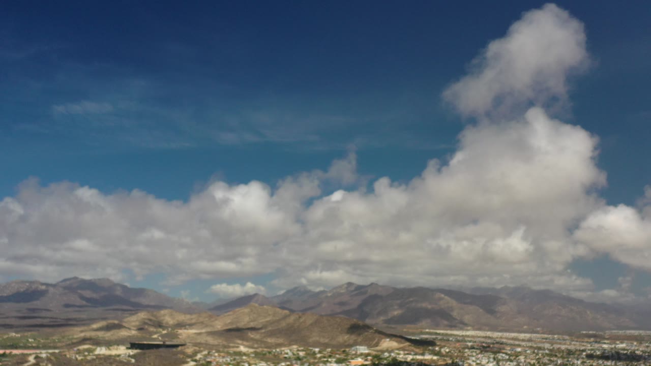 Opening frame of Los Cabos, Mexico from above, with town and trees low in frame, dry mountain ridges on the horizon, broad blue sky, and no visible people.