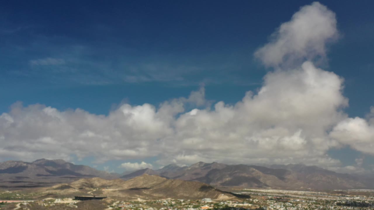 Los Cabos, Mexico aerial frame showing a large vertical cloud mass near the upper center, hazy mountain slopes beyond the town, and no visible people.