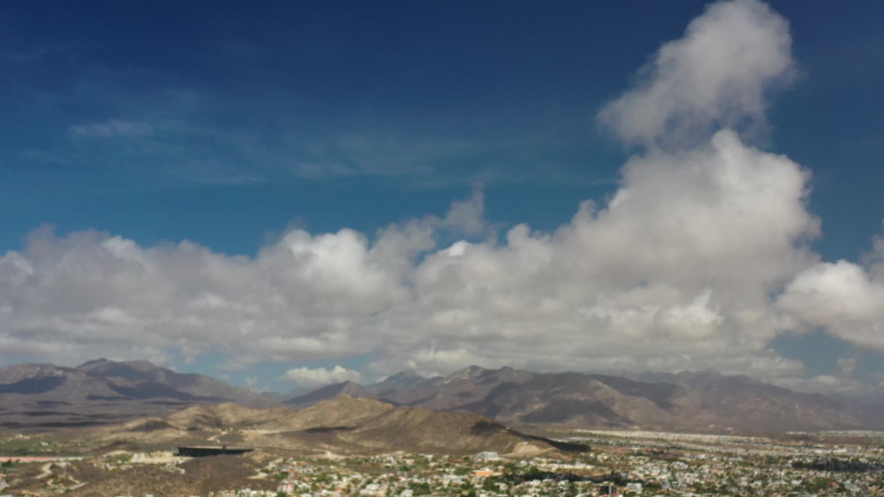 Aerial view across low hills, city blocks, mountain range, and scattered clouds.