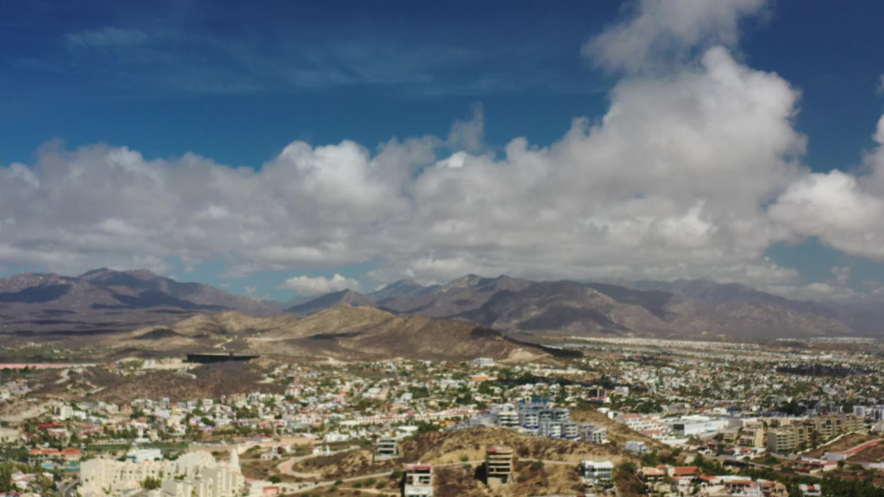 Opening frame of Los Cabos, Mexico, showing a wide cityscape beneath dry brown hills, distant mountains, blue sky, and large clouds, with no visible people.