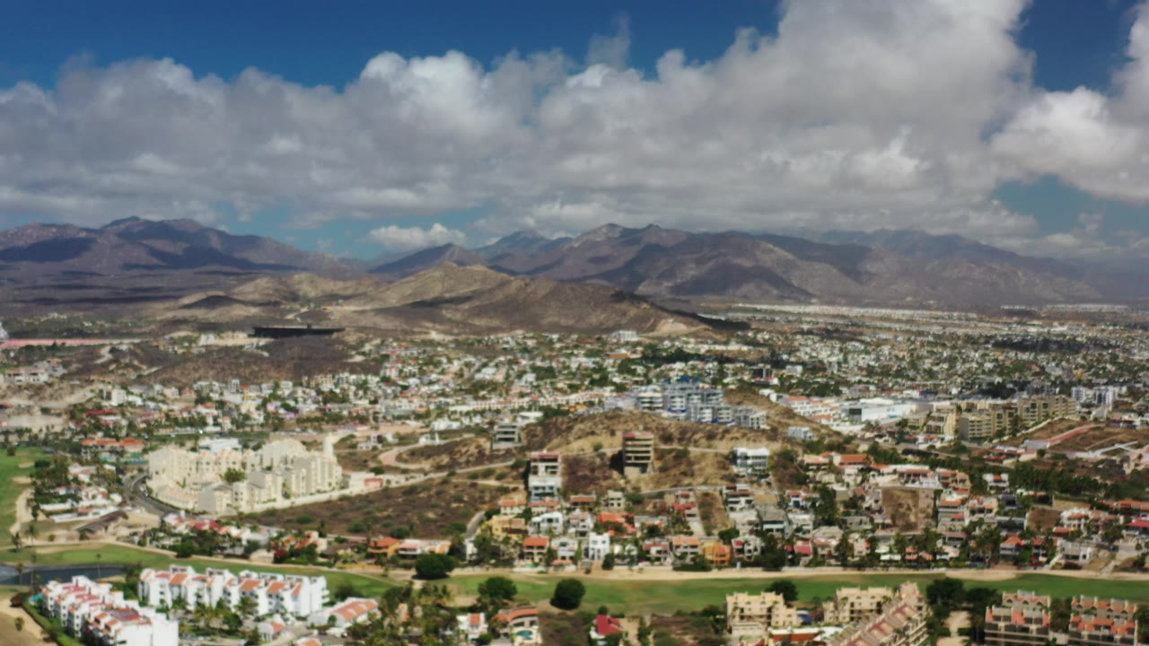 Wide aerial view of a Los Cabos town below dry mountains and clouds.